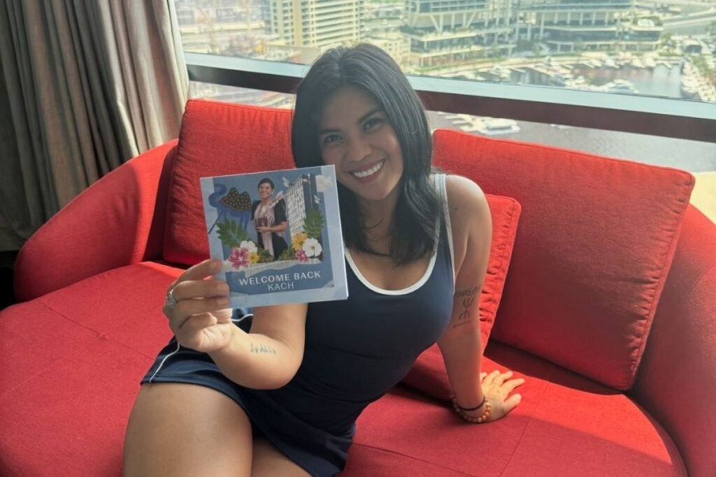 Kach smiling while sitting on a red couch, holding a