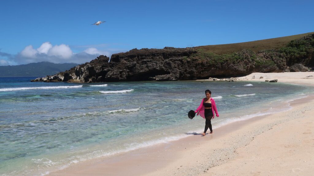 Woman walking along a white sand cove in Batanes with a rocky headland behind her and a small plane overhead